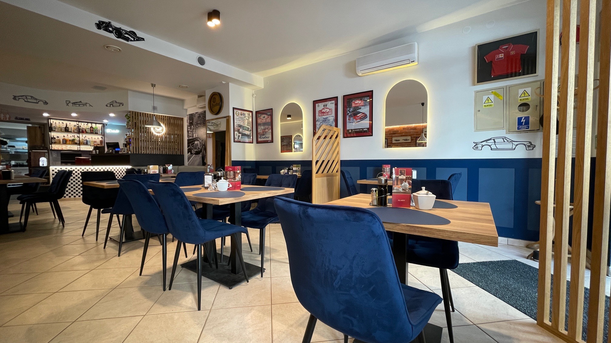 Wnętrze restauracji z motywem motoryzacyjnym: niebieskie krzesła, drewniane stoły z nakryciem, bar z tyłu, dekoracje ścienne nawiązujące do wyścigów samochodowych, w tym sylwetki aut i plakaty.