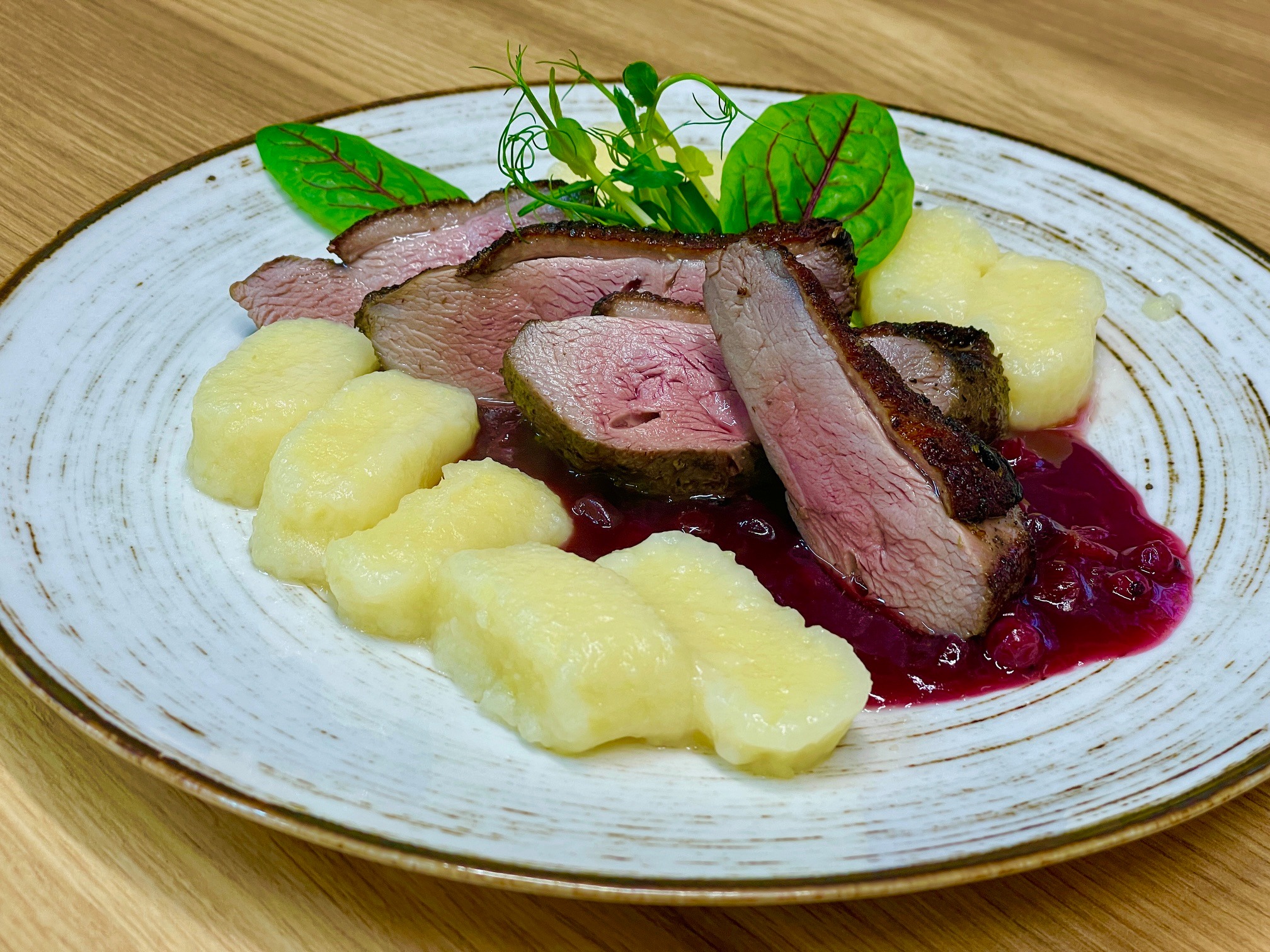 Elegancko podane plastry pieczonej kaczki z sosem żurawinowym i kluskami ziemniaczanymi, udekorowane świeżymi ziołami na talerzu z delikatnym wzorem.