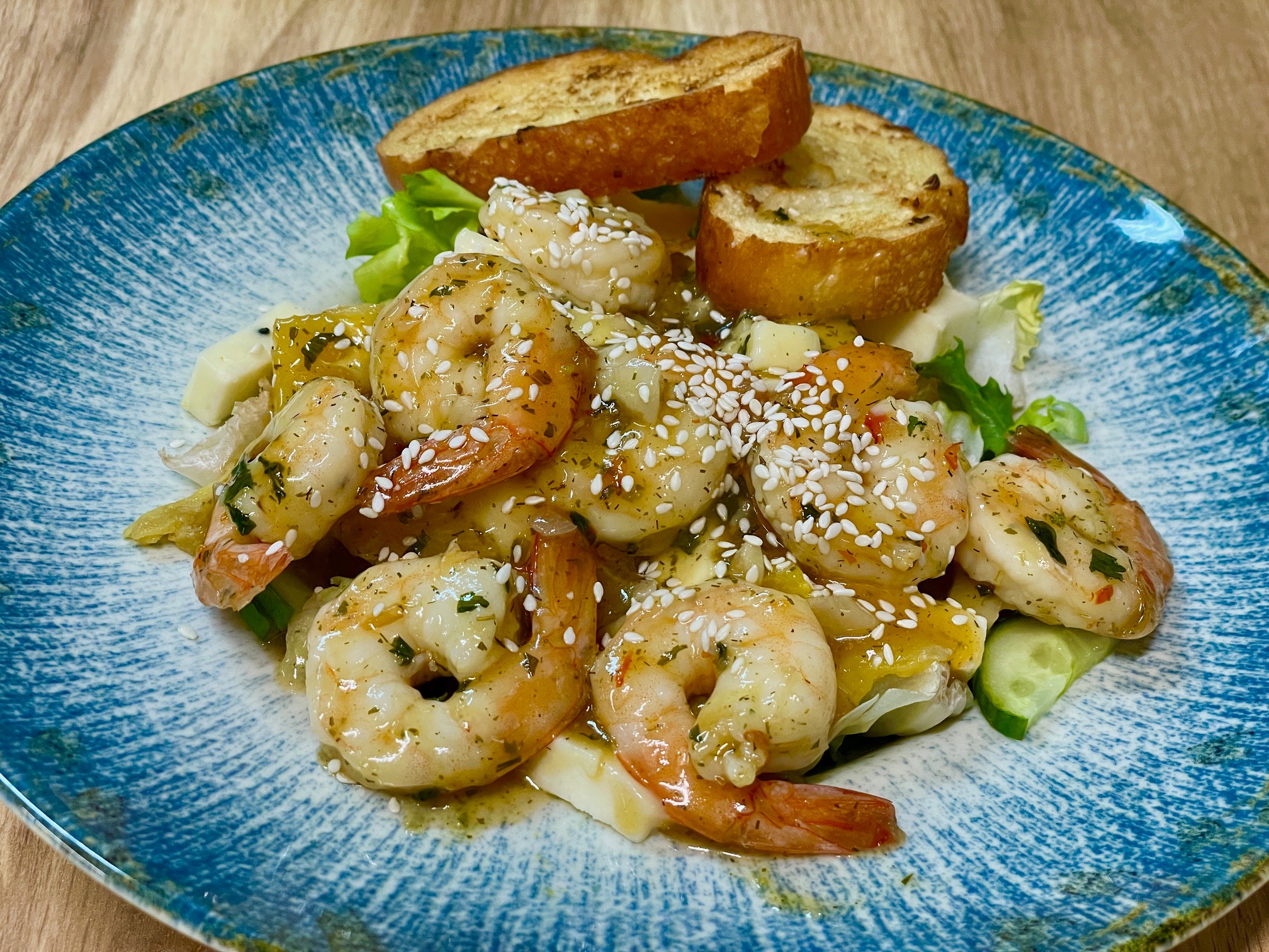 Apetycznie wyglądająca sałatka z krewetek z sezamem, grzankami i kawałkami warzyw na niebieskim talerzu z efektem marmuru, umieszczonym na drewnianym stole.