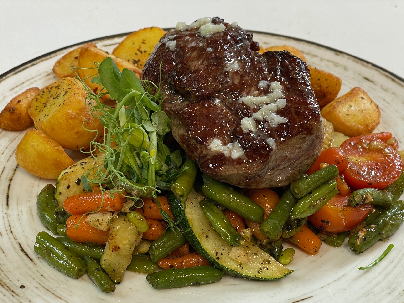 Soczysty stek wołowy z serem, podany na talerzu z pieczonymi ziemniakami i warzywami: fasolką szparagową, marchewką, cukinią i pomidorami.