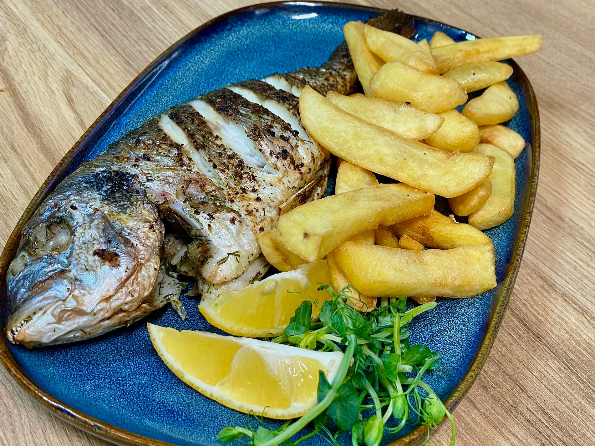 Grilowana ryba dorada z frytkami, plastrami cytryny i świeżymi ziołami na niebieskim talerzu z brązowym brzegiem, ułożona na drewnianym stole.