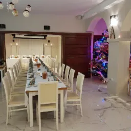 Długi stół z białym obrusem i kremowymi krzesłami, nakryty do przyjęcia, z napojami w dzbankach, w tle choinka i dekoracje świąteczne w przestronnym, jasnym wnętrzu.