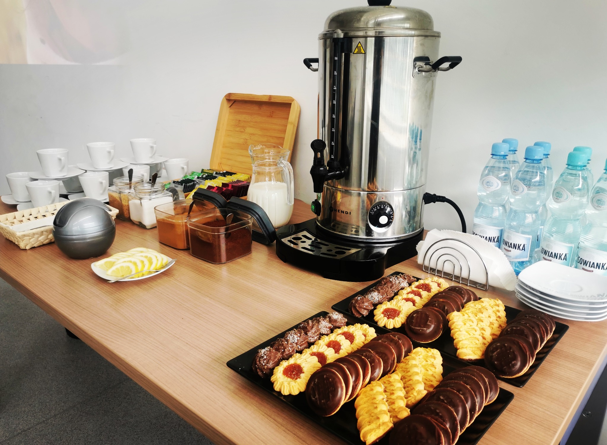 Stół z przekąskami: ciasteczka na czarnych talerzach, plastry cytryny, dzbanek mleka, zapas herbat, cukier, woda butelkowana i podgrzewacz do wody Hendi.