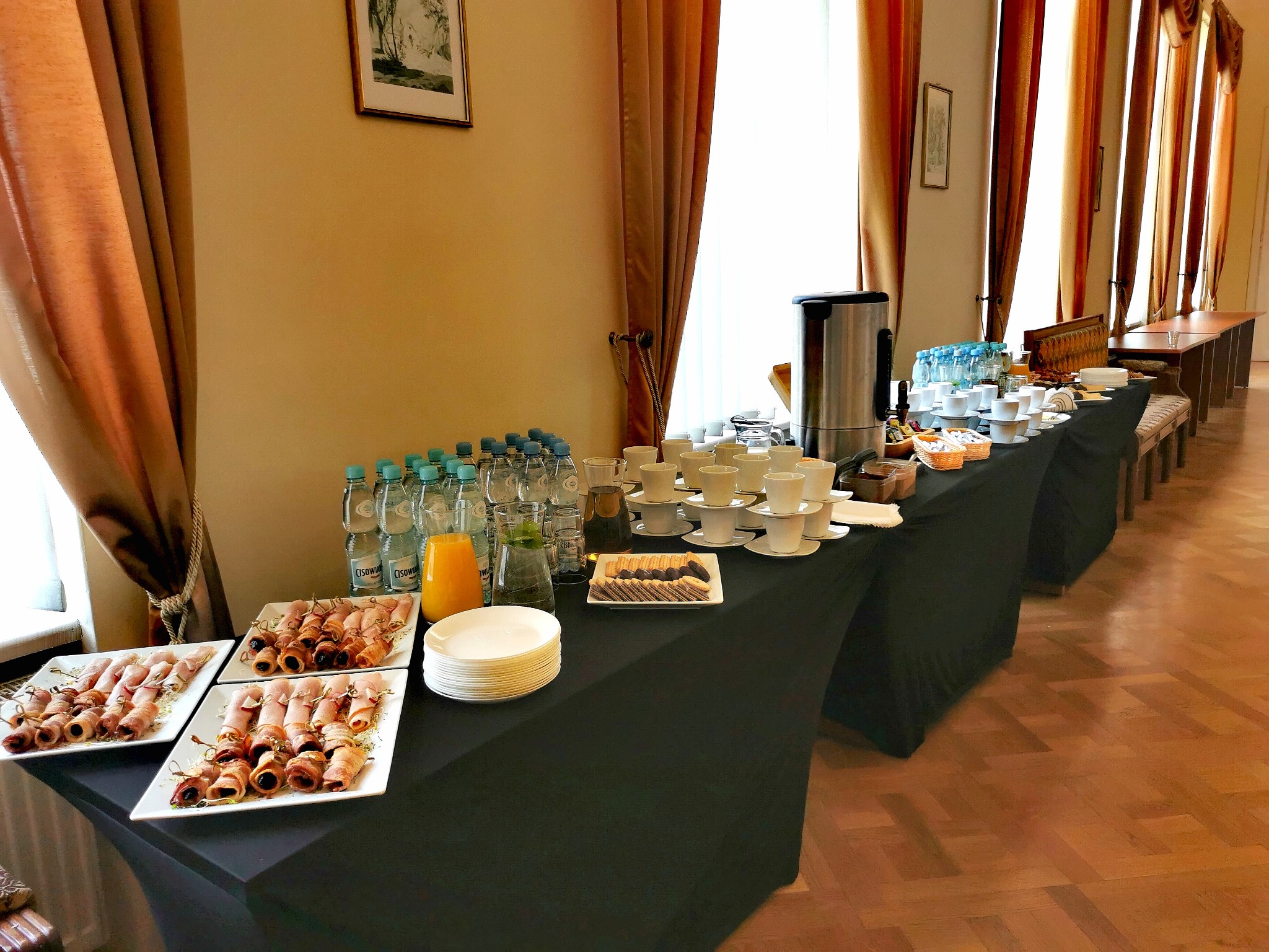 Stół z przekąskami, napojami i kawą podczas wydarzenia firmowego, widoczne koreczki z szynki i śliwki, woda w butelkach, dzbanek soku pomarańczowego, ciastka i zastawa stołowa.