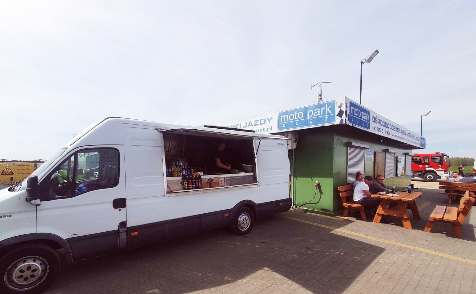 Biały food truck z otwartym oknem sprzedaży, zaparkowany obok zielonego budynku Moto Parku z klientami siedzącymi przy drewnianych stołach piknikowych. W tle widać wóz strażacki.