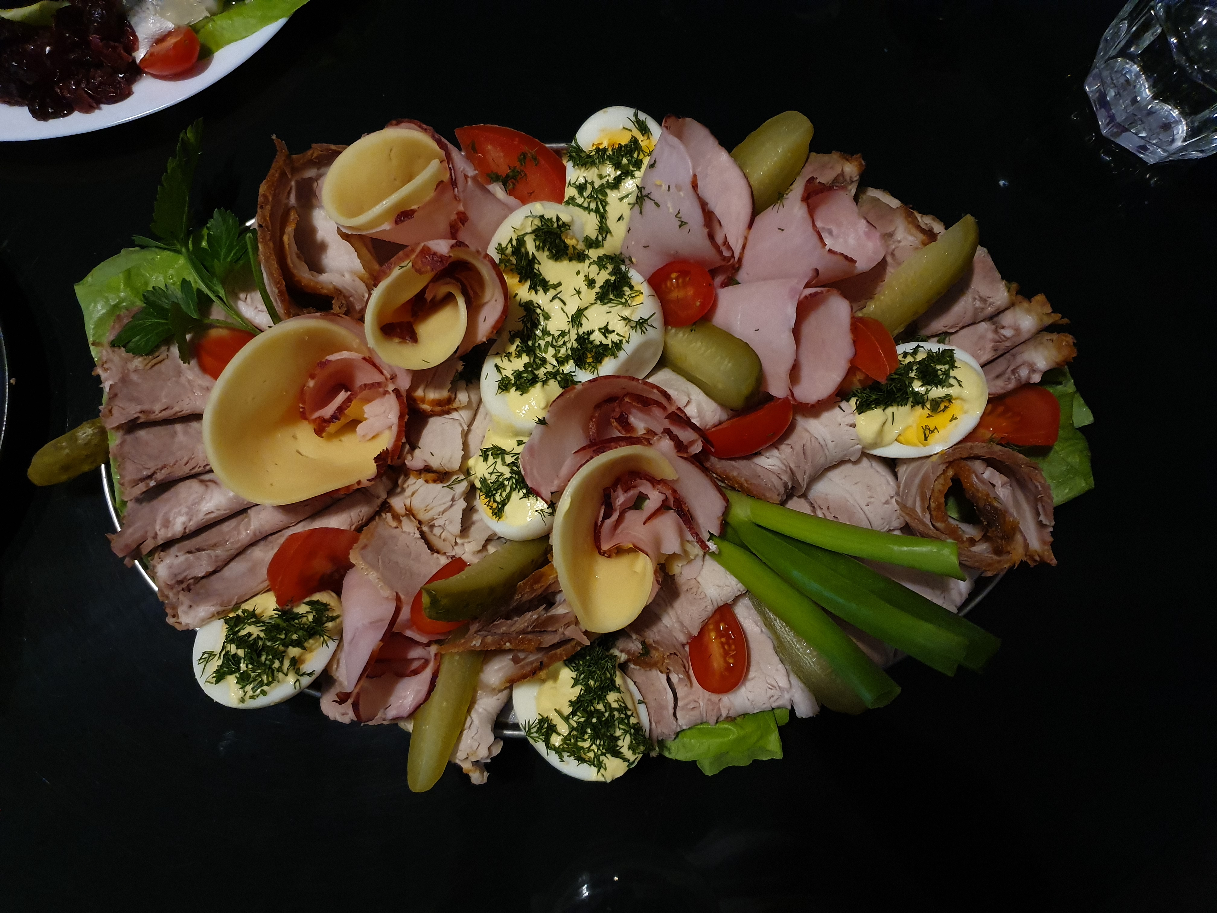 Elegancka taca z wędlinami, jajkami w majonezie posypanymi koperkiem, roladkami z szynki i sera oraz świeżymi warzywami (pomidorki koktajlowe, ogórki kiszone, zielona cebulka), ułożona na czarnym...