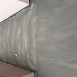 Świeżo pomalowana, szara ściana balkonu z widocznymi śladami pędzla, z pozostawioną roboczą rękawicą i częściowo wykończoną podłogą z korka.