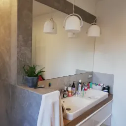 Nowoczesna łazienka z umywalką nablatową, białymi szafkami z drewnianym blatem, dużym lustrem w szarej ramie, wiszącymi lampami i rośliną w doniczce