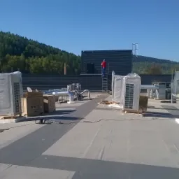 Instalacja systemów na dachu budynku: jednostki zewnętrzne pod folią, kanały wentylacyjne, pracownik na drabinie przy czarnej elewacji na tle zalesionych wzgórz.