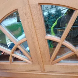 Fragment dębowych drzwi zewnętrznych z półokrągłym przeszkleniem, widoczne prace wykończeniowe, malowanie czerwoną farbą.