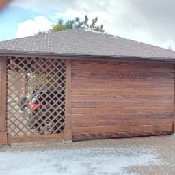 Drewniana wiata garażowa z kratownicą i brązowym gontem bitumicznym, częściowo zasłaniająca samochód, na tle zimowego krajobrazu.