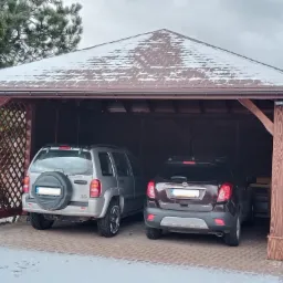 Zadaszone miejsce parkingowe z dwoma samochodami, dach pokryty brązowym gontem bitumicznym z resztkami śniegu, drewniane słupy i ażurowa ścianka boczna.