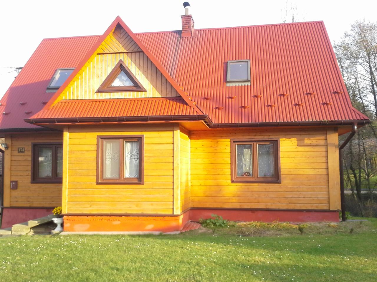 Żółty dom z czerwonym dachem z blachy falistej, trójkątne okno na poddaszu, widok z trawnika w słoneczny dzień.