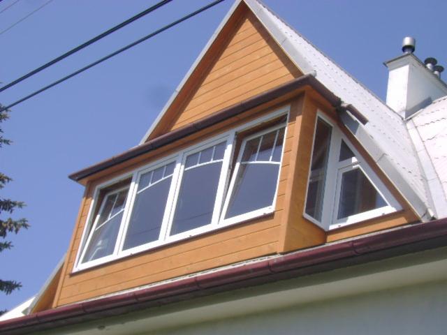 Lukarna z białymi oknami, drewnianą elewacją i metalowym dachem na tle błękitnego nieba.