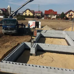 Wznoszenie fundamentów z bloczków betonowych na piaszczystym podłożu, widoczny betonowóz z wysięgnikiem, szalunki oraz teren budowy z domami w tle.