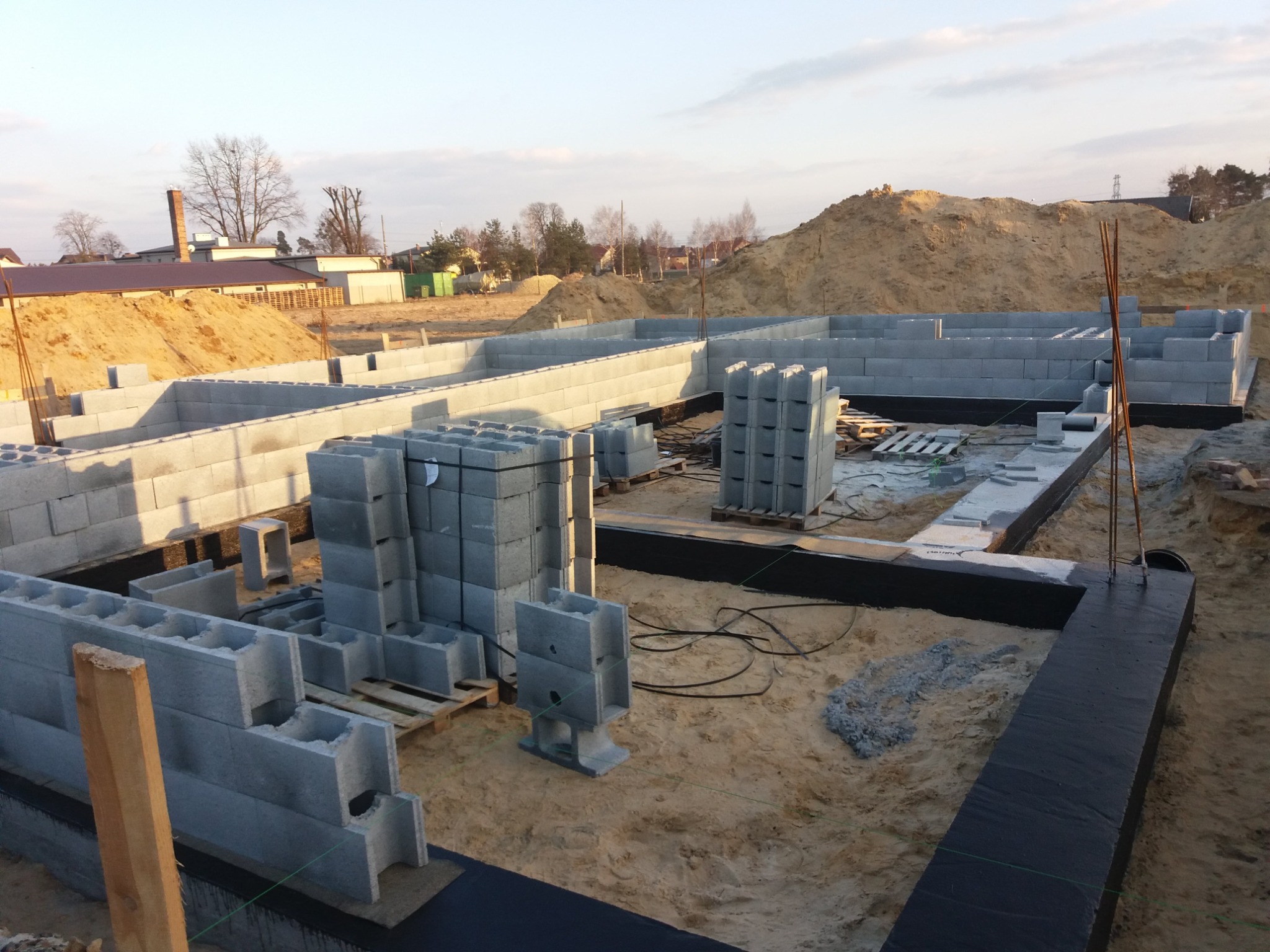 Wznoszenie ścian fundamentowych z bloczków betonowych na warstwie hydroizolacji, widoczne zbrojenie pionowe i zapas bloczków na paletach, w tle usypane pryzmy ziemi.