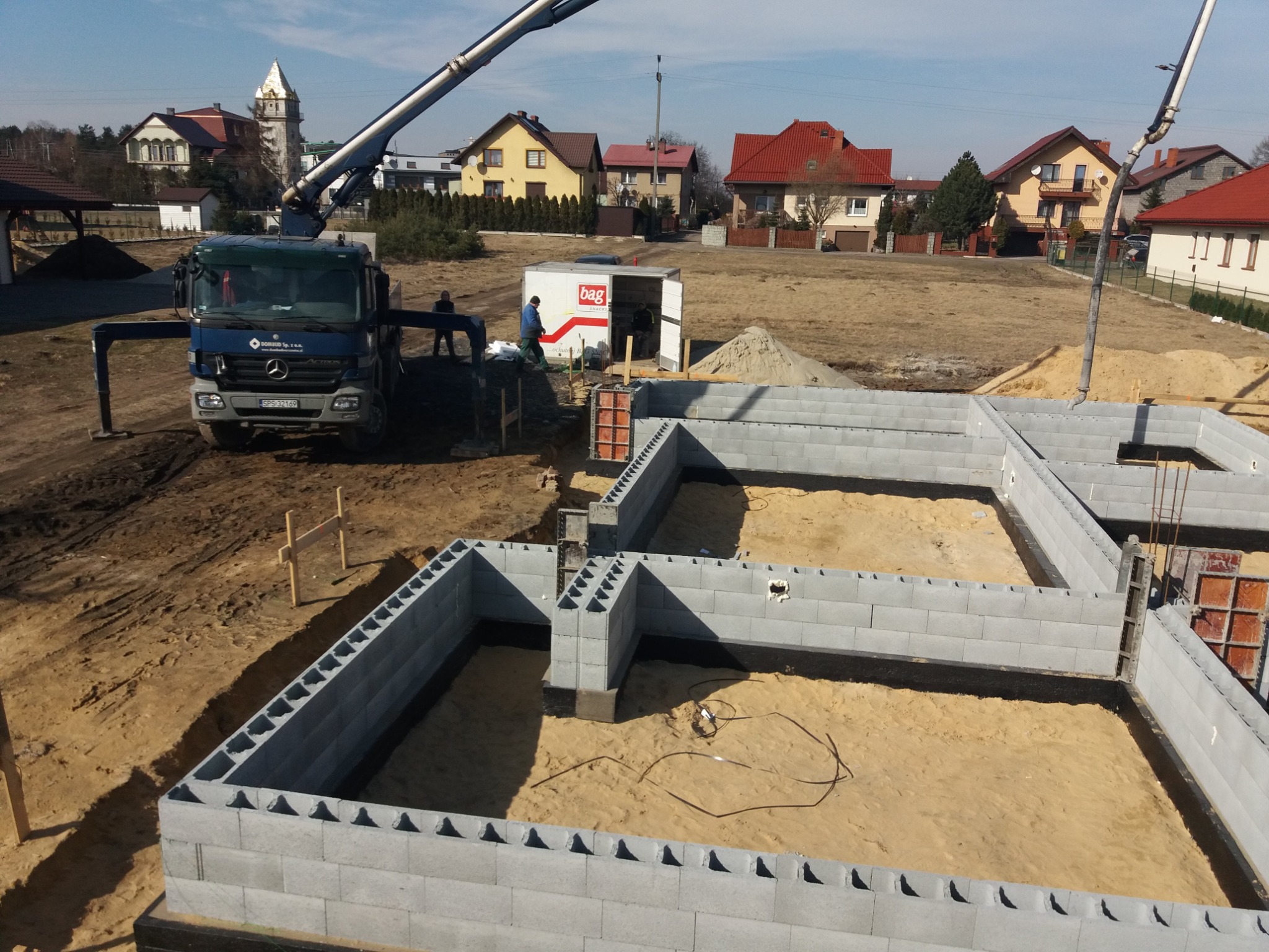 Wznoszenie fundamentów z bloczków betonowych na piaszczystym podłożu, widoczny betonowóz z wysięgnikiem, szalunki oraz teren budowy z domami w tle.
