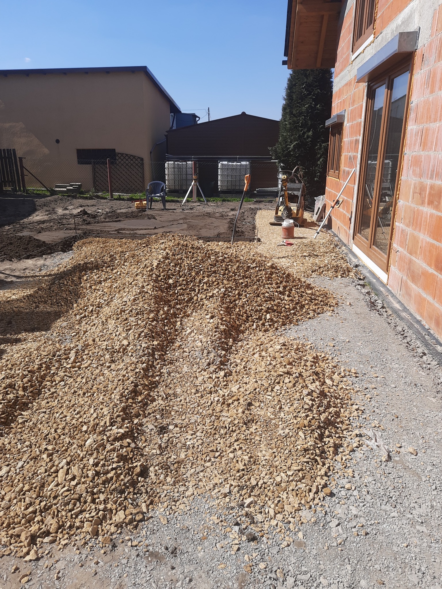 Stos żółtego kruszywa obok ściany budynku z pustaków ceramicznych, przygotowany teren pod budowę, zagęszczarka w tle.