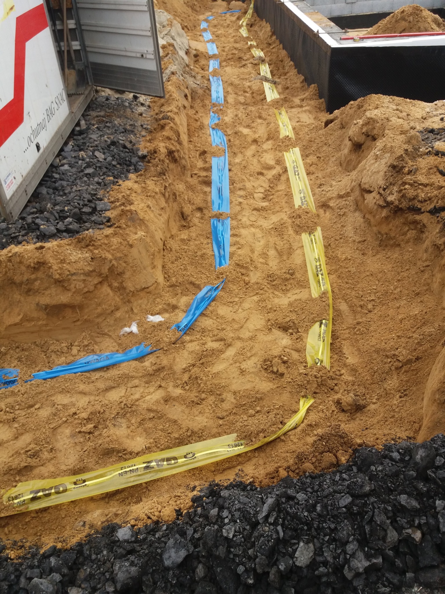 Wykop w Rudziczce z widocznymi taśmami ostrzegawczymi w kolorze żółtym i niebieskim, oznaczającymi przebieg instalacji, obok fundamentów budynku i hałdy ziemi.