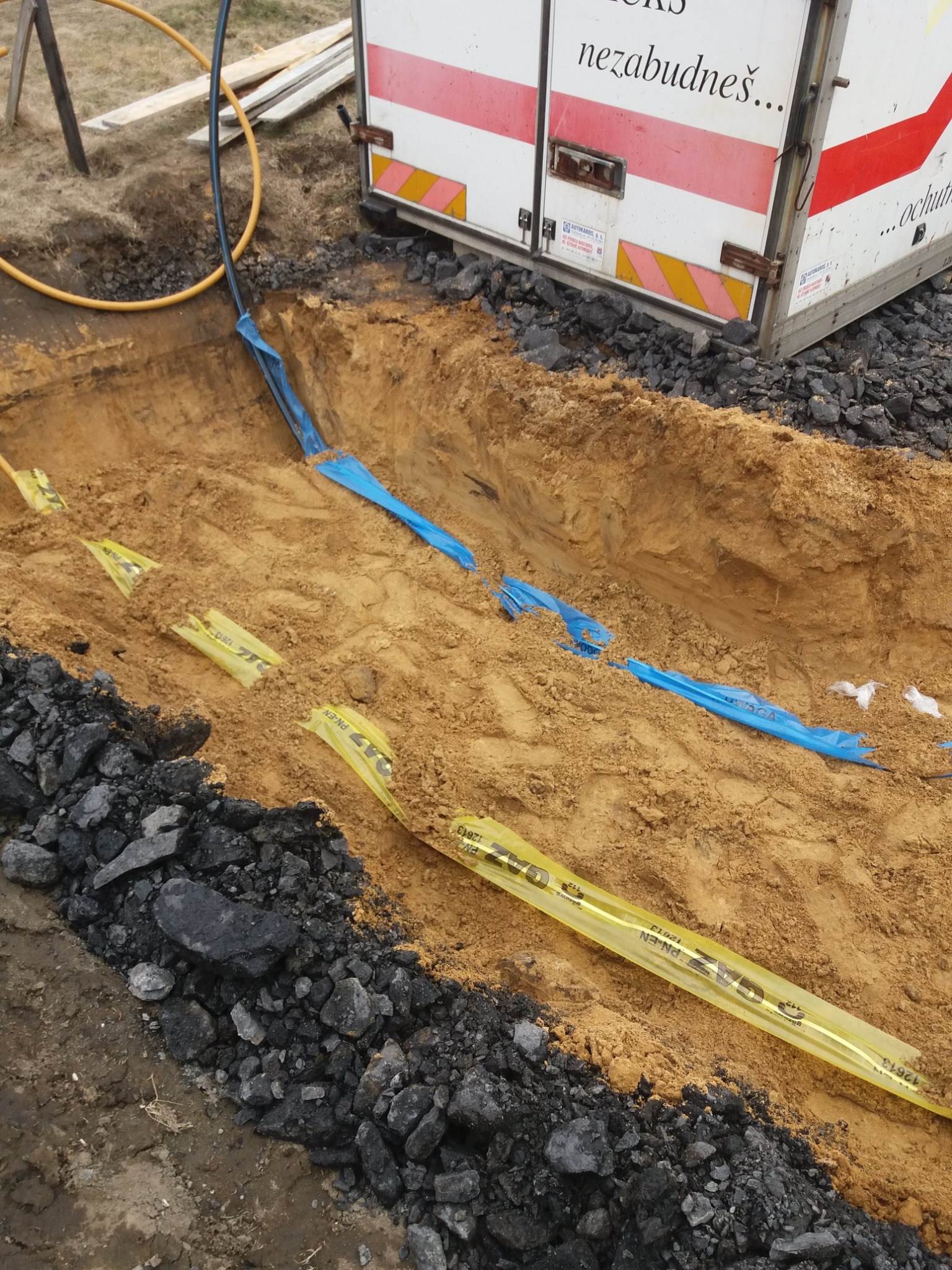 Wykop w ziemi z widocznymi rurami gazowymi oznaczonymi żółtą taśmą ostrzegawczą oraz niebieską folią ochronną, obok leży pryzma czarnego gruzu, w tle fragment samochodu dostawczego z reklamą.
