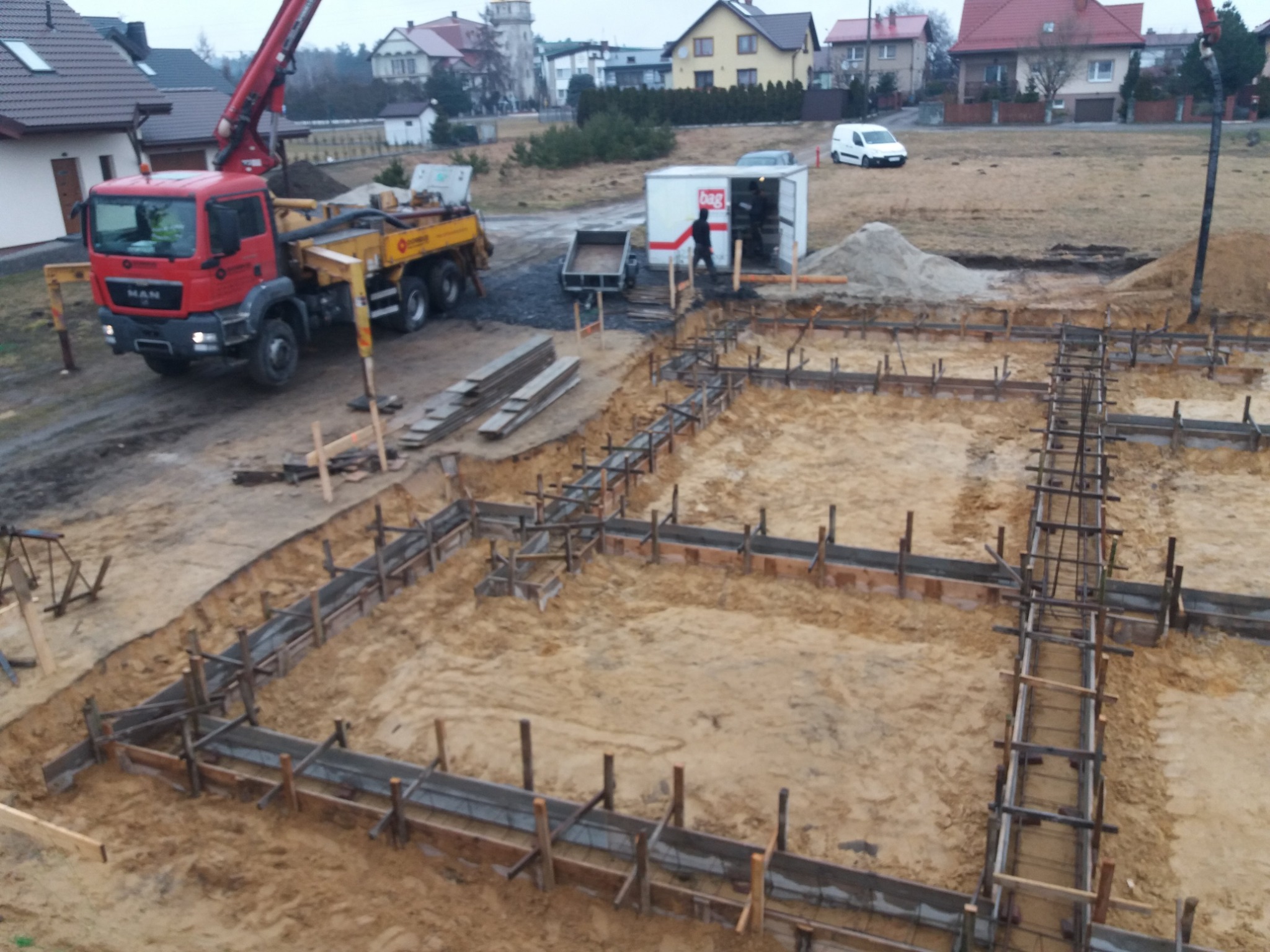 Przygotowanie szalunków i zbrojenia pod fundamenty domu jednorodzinnego, widoczna pompa do betonu MAN i zaplecze budowy w tle.