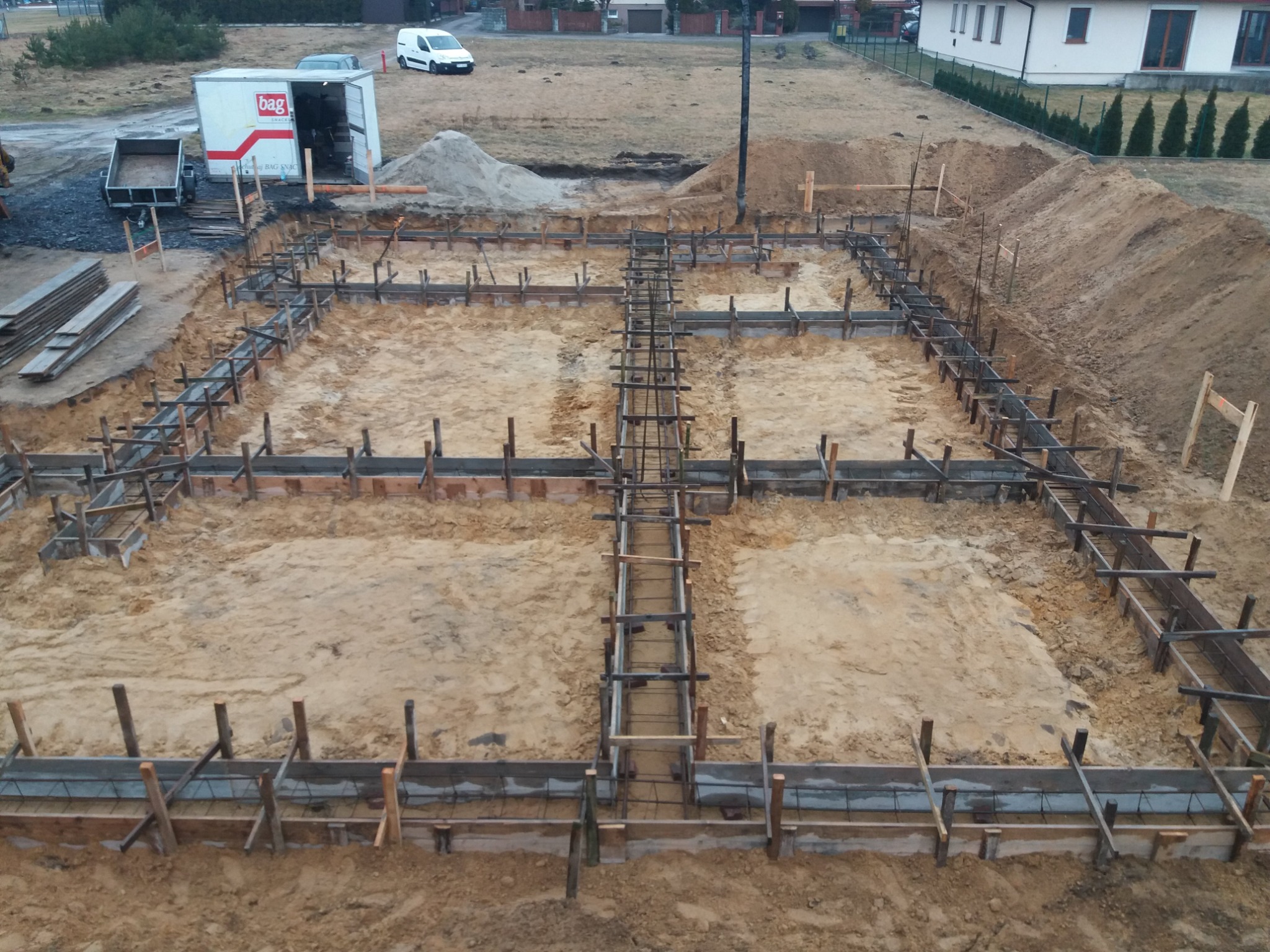 Szeroki kadr z góry przedstawia przygotowany szalunek i zbrojenie pod fundamenty budynku na placu budowy, widoczny wykop w ziemi, z tyłu zaparkowany biały van i przyczepa.