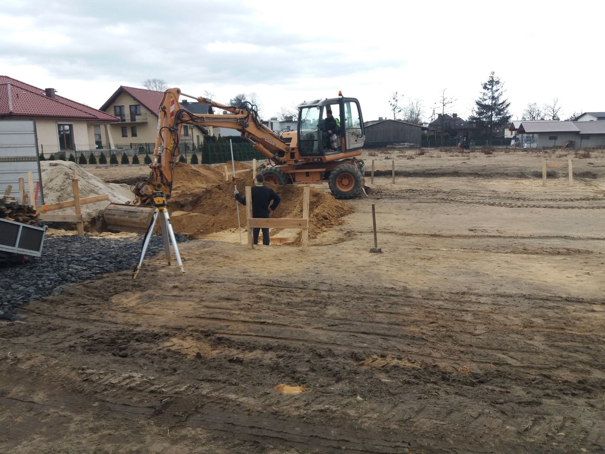 Żółta koparka Case na placu budowy, operator w kabinie, pracownik mierzy poziom gruntu za pomocą łaty niwelacyjnej, widoczny niwelator laserowy na trójnogu, wykop pod fundamenty, drewniane szalunki...