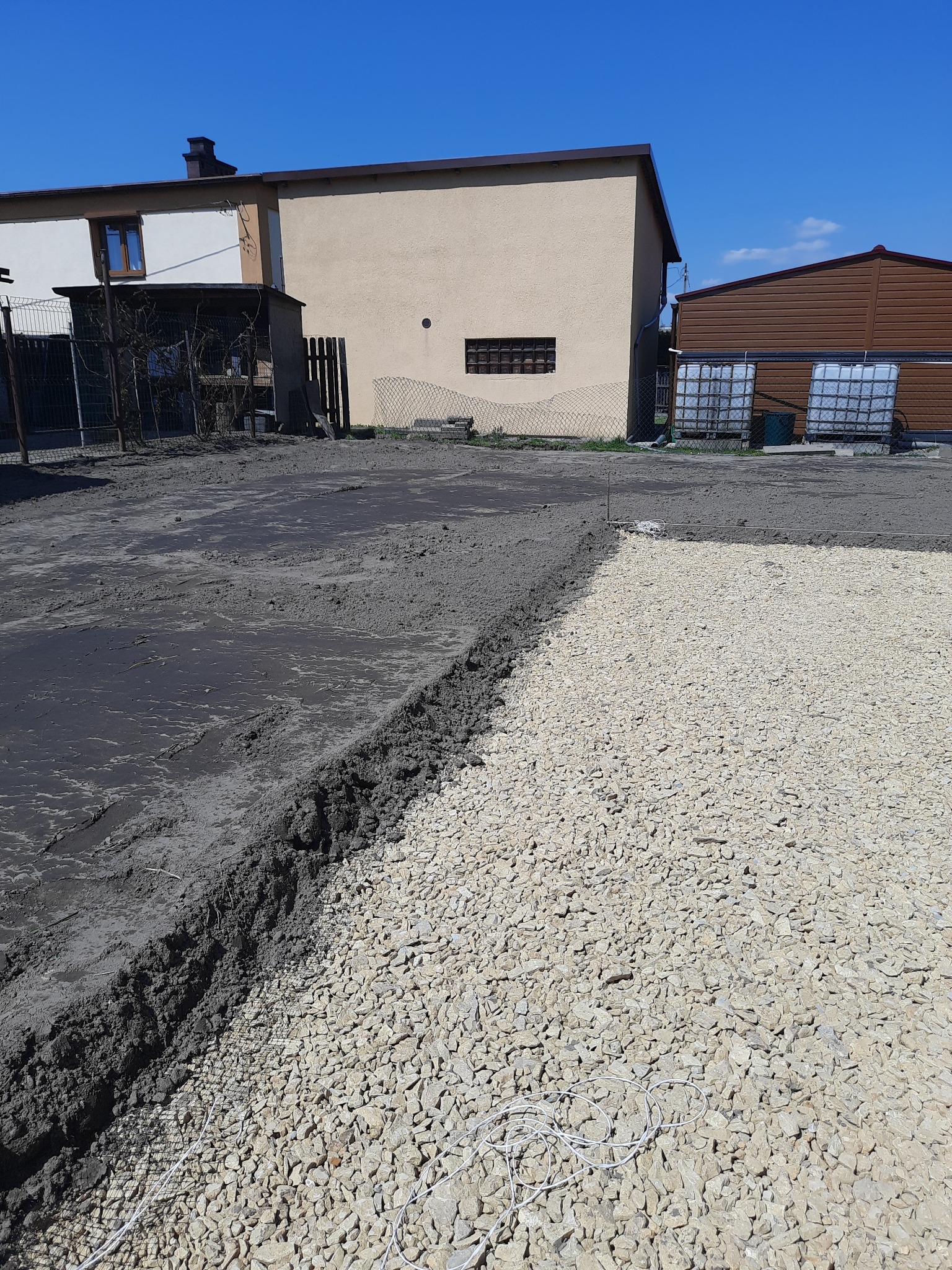 Przygotowanie podłoża pod nawierzchnię: warstwa ubitego kruszywa sąsiaduje z warstwą mokrej, ciemnej ziemi. W tle budynki gospodarcze i błękitne niebo.