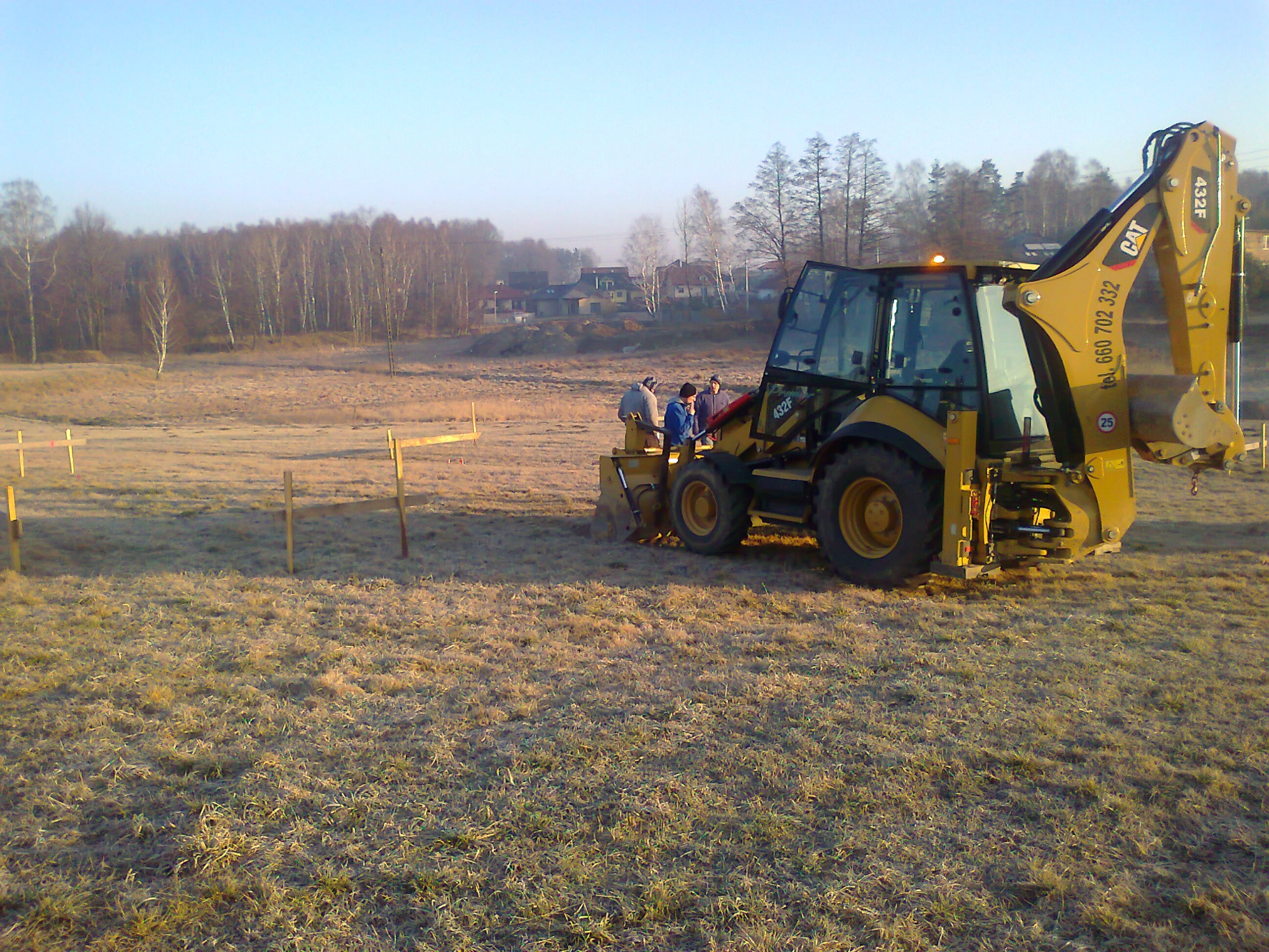 Żółta koparko-ładowarka CAT 432F na zaśnieżonym polu, grupa osób w pobliżu kabiny, drewniane paliki wbite w ziemię, w tle zabudowania i drzewa.