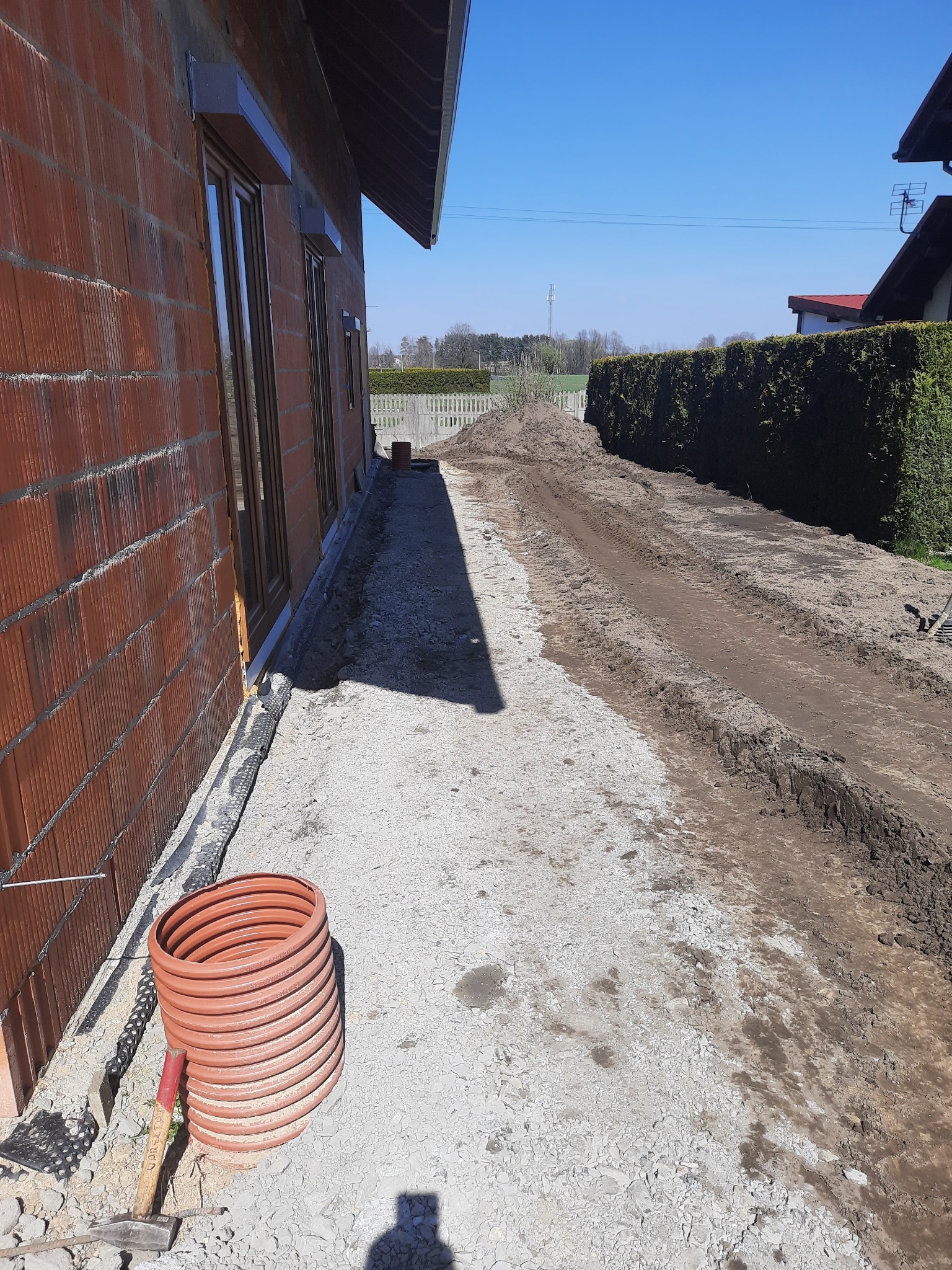 Przygotowanie terenu pod budowę domu: widoczny fragment ściany z czerwonej cegły, zwój rur drenarskich przy ścianie, ubity tłuczeń i ślady opon po ciężkim sprzęcie na ziemi, w tle usypana ziemia...