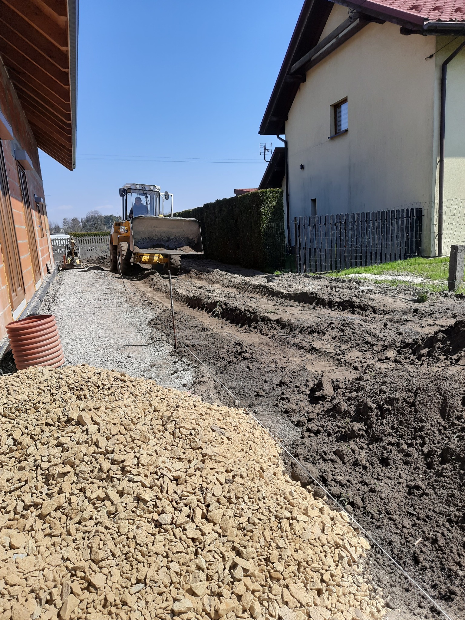 Żółta koparko-ładowarka wyrównuje teren pod budowę, widoczny stos kruszywa, rury kanalizacyjne i częściowo utwardzona droga w słoneczny dzień.