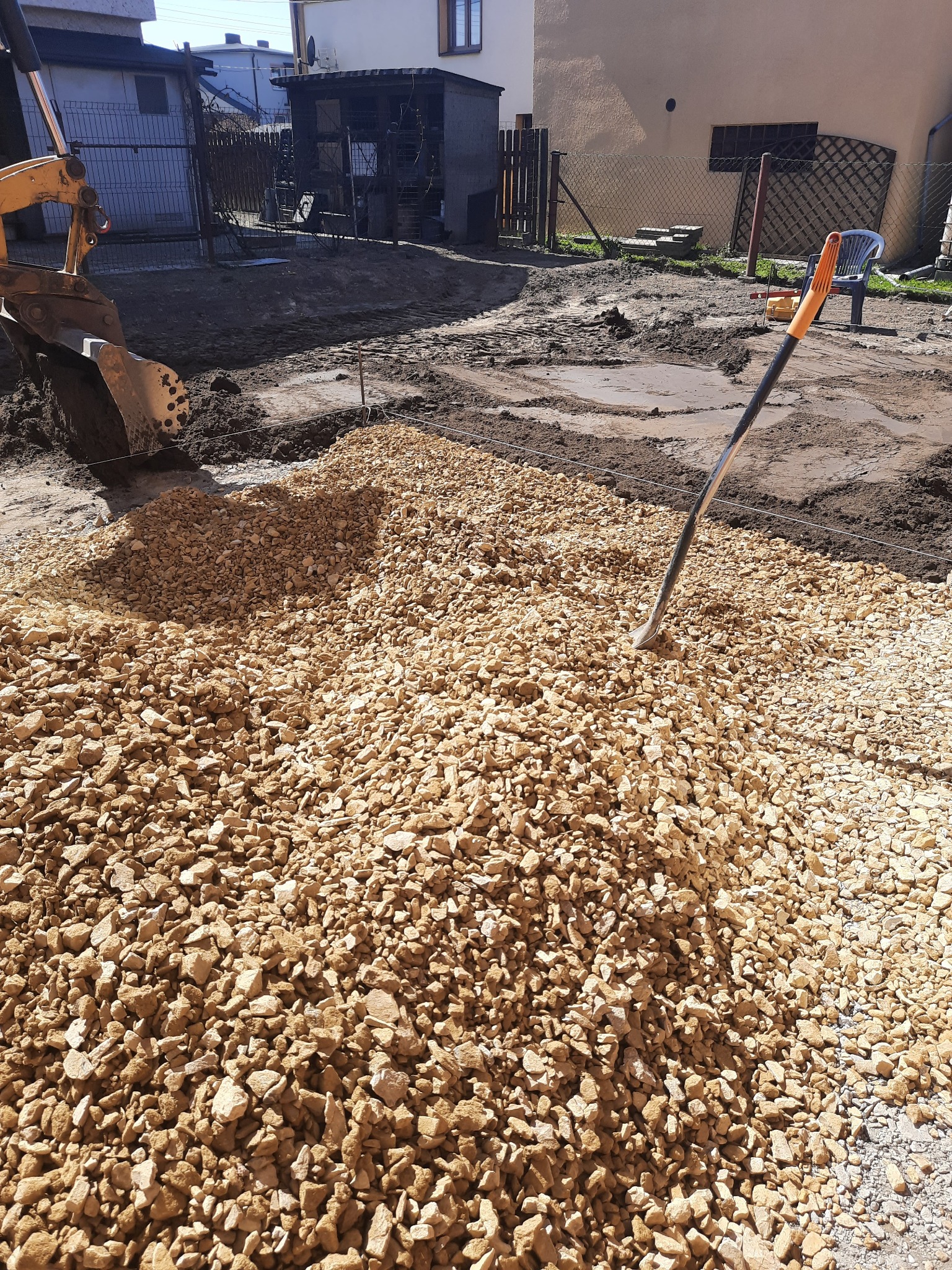 Żółty kruszywo wysypane na przygotowany teren podłoża, widoczna łyżka koparki i szpadel wbity w kruszywo, w tle teren budowy z domami i ogrodzeniem.