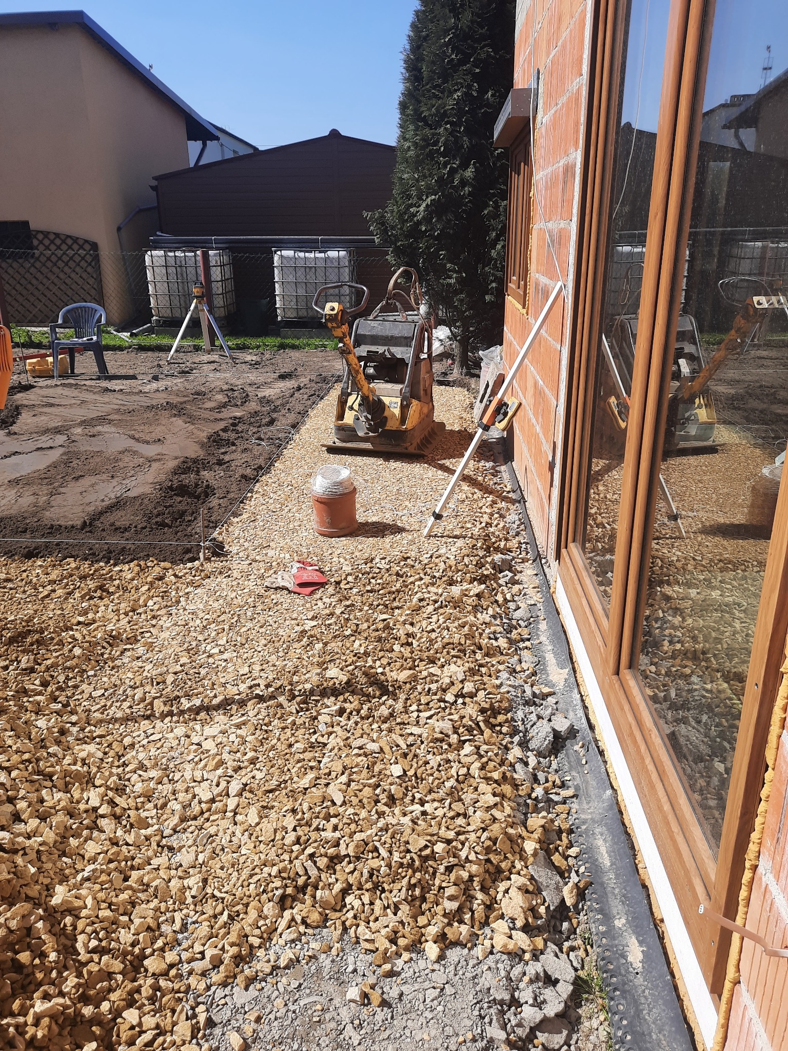 Przygotowanie podłoża pod kostkę brukową: wyrównany teren z podsypką żwirową, zagęszczarka w tle, widoczne narzędzia i budynek w stanie surowym otwartym.