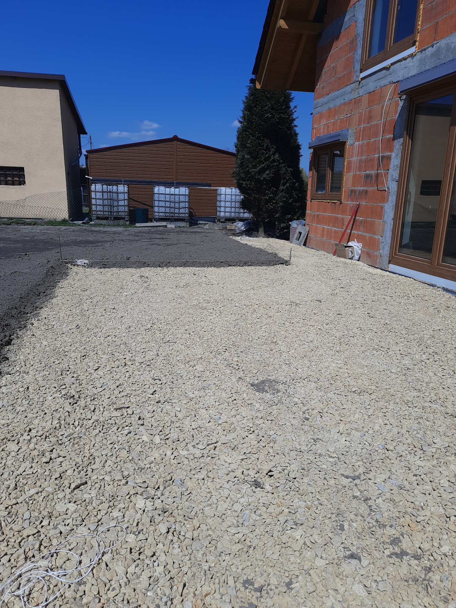 Przygotowanie terenu pod budowę: warstwa utwardzająca z jasnego kruszywa, częściowo zalana betonem, widok na budynek w trakcie wznoszenia z czerwonej cegły i zabudowania gospodarcze w tle.