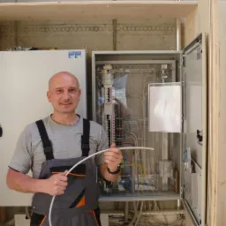 Elektryk w szarym kombinezonie trzyma biały kabel na tle otwartej szafy rozdzielczej z okablowaniem i bezpiecznikami, zamontowanej w drewnianej obudowie.