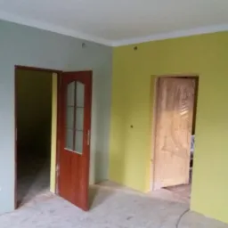 Wnętrze po odświeżeniu: ściany w odcieniach żółci i szarości, widoczne dwie ościeżnice z drzwiami, jedne z nich osłonięte folią ochronną.