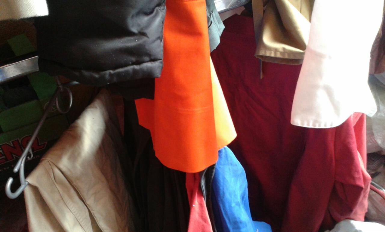 Różnokolorowe ubrania używane wiszące na metalowym wieszaku, w tym pomarańczowa kurtka i beżowa koszula, w nieostrym świetle.