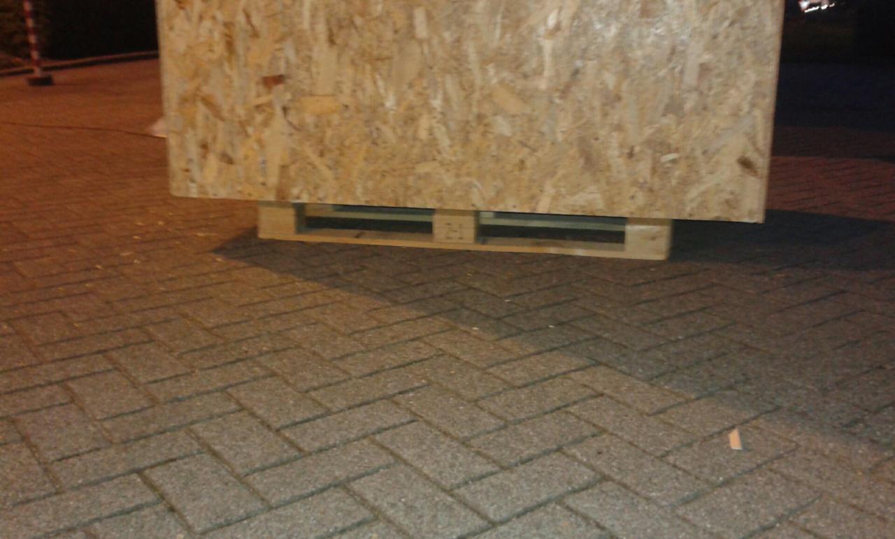 Duża drewniana skrzynia z płyty OSB umieszczona na palecie, stojąca na brukowanym podłożu w słabym oświetleniu.