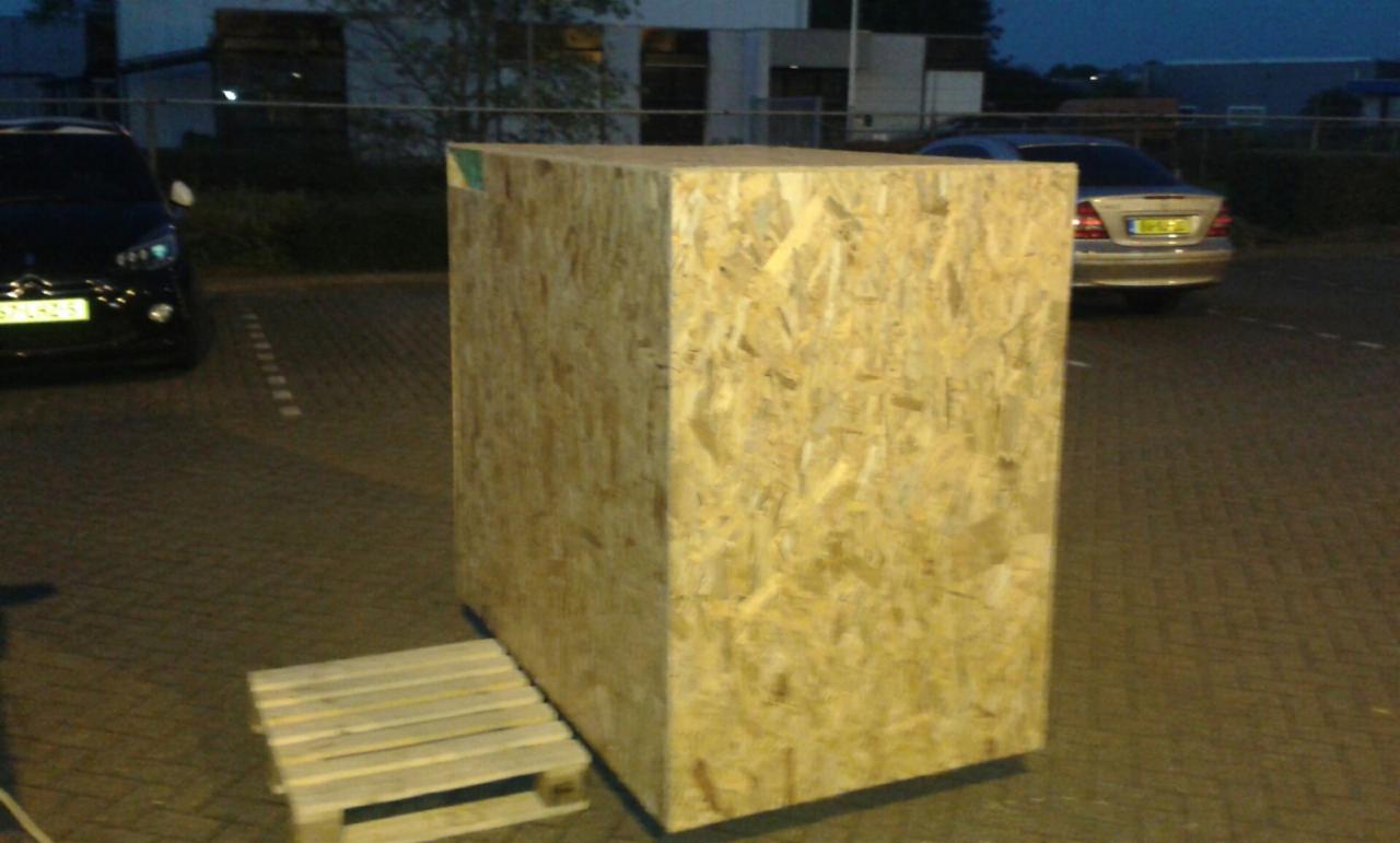 Duża skrzynia z płyty OSB na palecie, zaparkowane samochody w tle, oświetlenie uliczne.