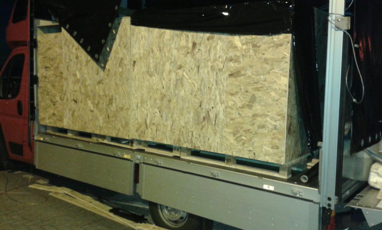 Duża, drewniana skrzynia z płyty OSB zabezpieczona czarną plandeką, umieszczona na palecie w przestrzeni ładunkowej czerwonego samochodu dostawczego.