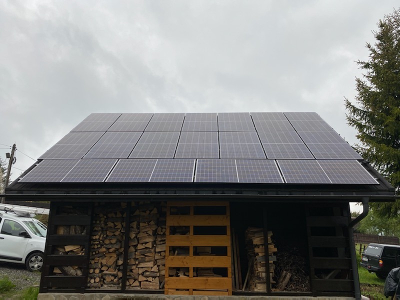 Instalacja paneli słonecznych na dachu budynku gospodarczego z drewnem opałowym w Piekarach Śląskich, widok z poziomu gruntu, z boku zaparkowany biały samochód dostawczy.