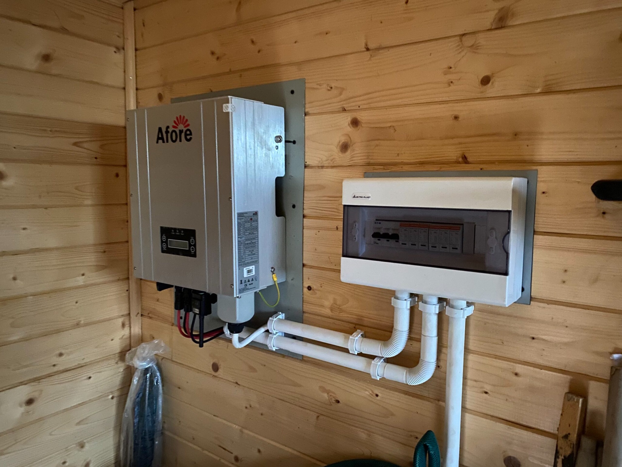 Szary falownik z logo Afore zamontowany na drewnianej ścianie obok skrzynki elektrycznej z widocznymi przewodami i rurami. Instalacja wewnątrz budynku.