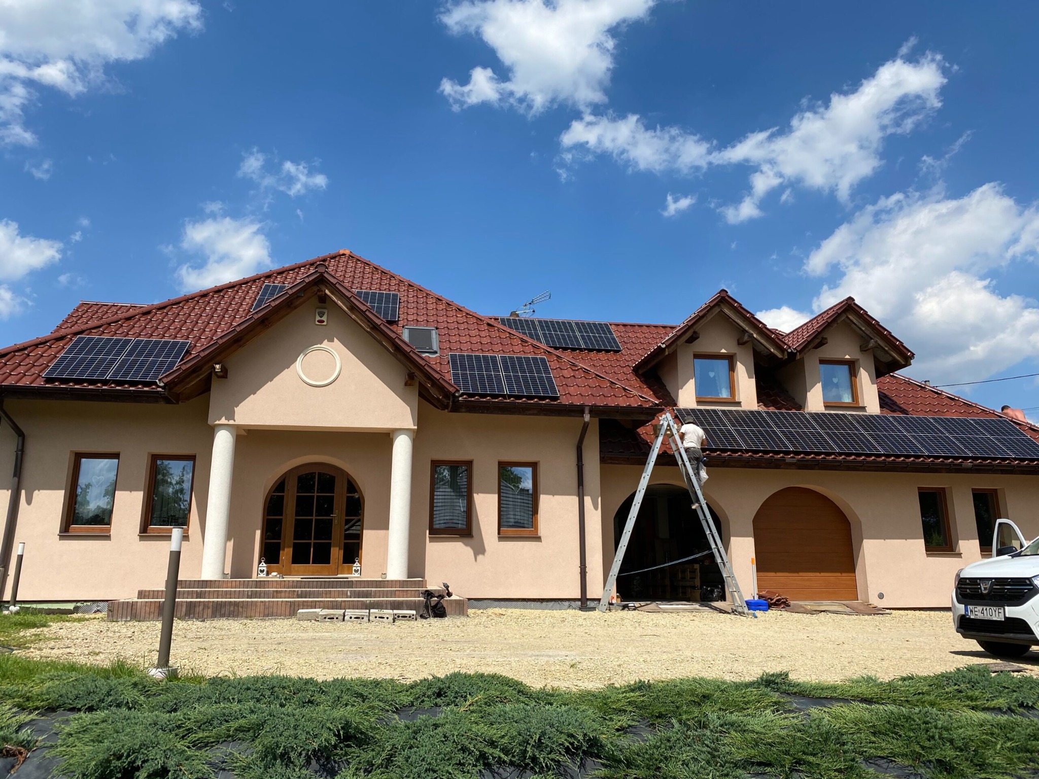 Montaż paneli słonecznych na dachu domu jednorodzinnego z czerwoną dachówką, pracownik na drabinie, błękitne niebo z chmurami w tle.