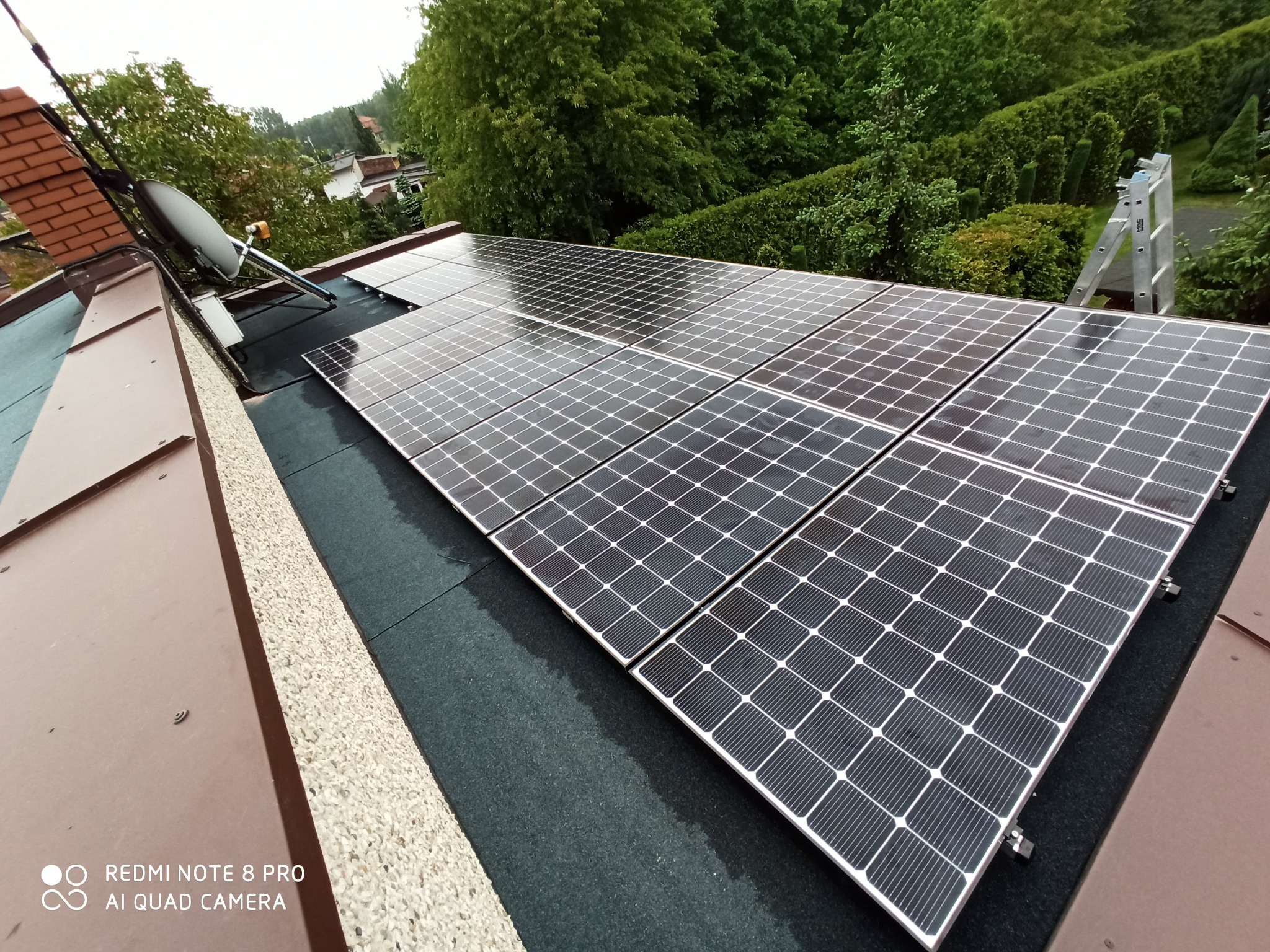 Instalacja paneli słonecznych na płaskim dachu pokrytym czarną membraną, widoczna antena satelitarna, drabina oparta o dach i zieleń w tle.