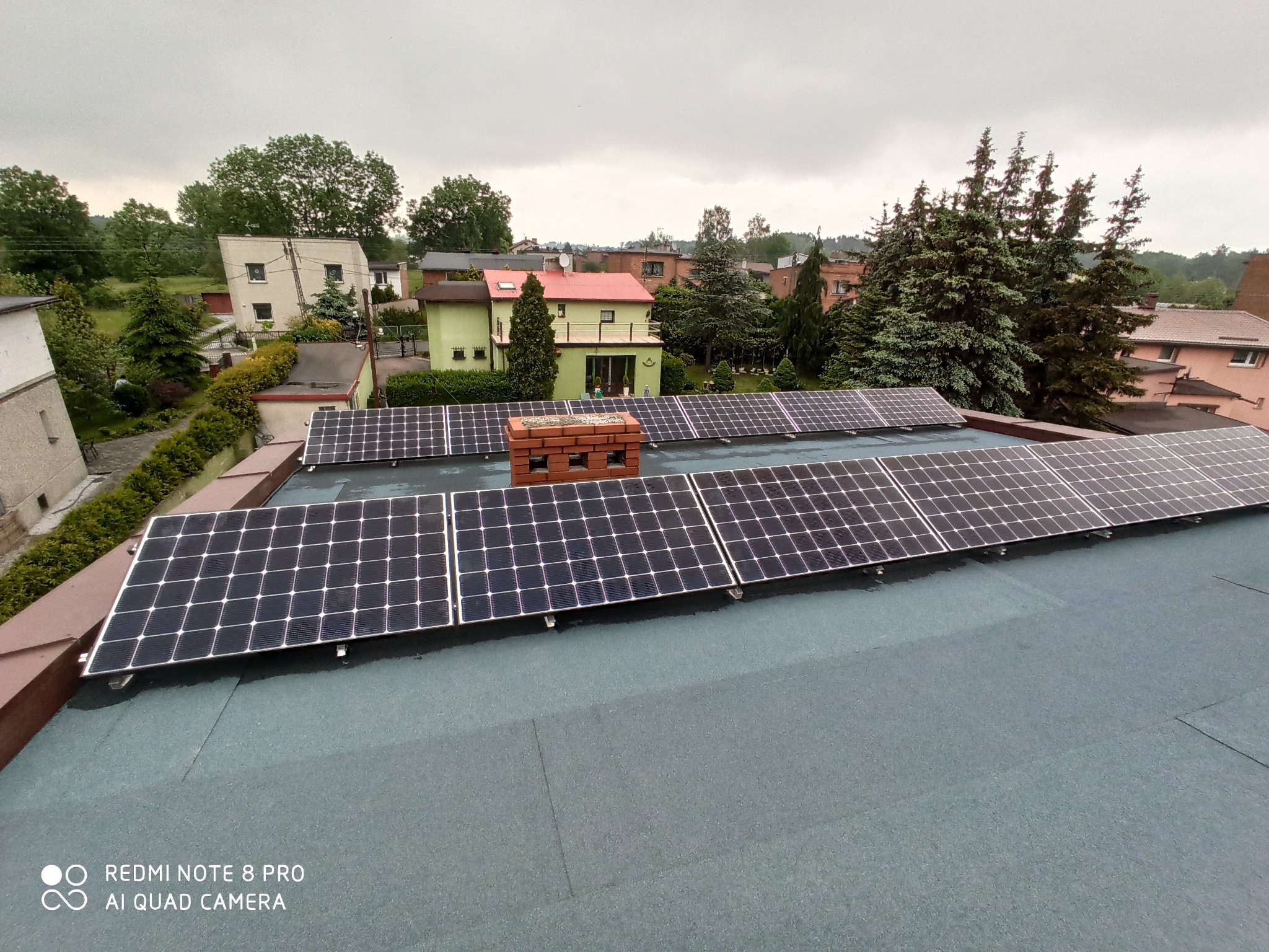 Rząd paneli słonecznych na płaskim dachu budynku mieszkalnego, z ceglanym kominem w tle i widokiem na okoliczną zabudowę w pochmurny dzień.