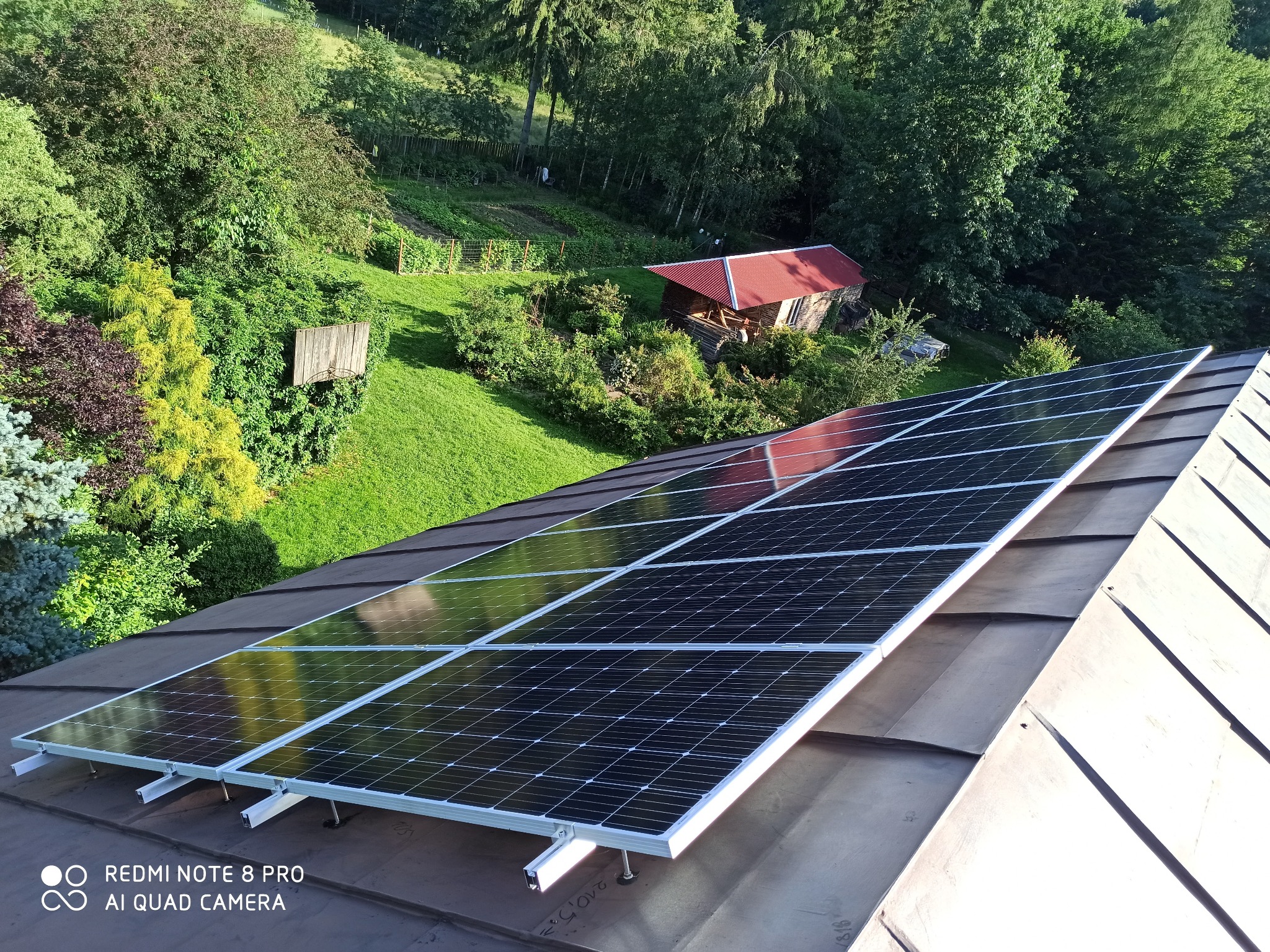 Panel słoneczny na dachu domu z widokiem na ogród i mały drewniany budynek w tle.