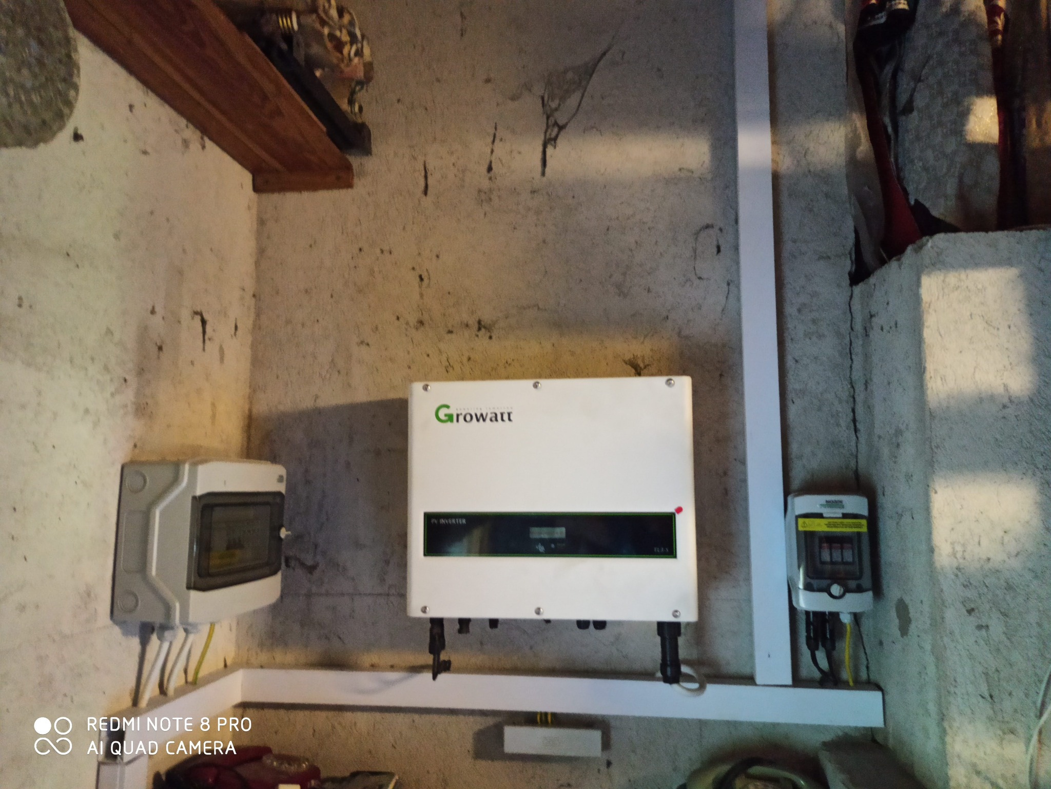 Biały inwerter Growatt PV zamontowany na szarej, surowej ścianie wewnątrz budynku, z widoczną skrzynką elektryczną i elementami instalacji.