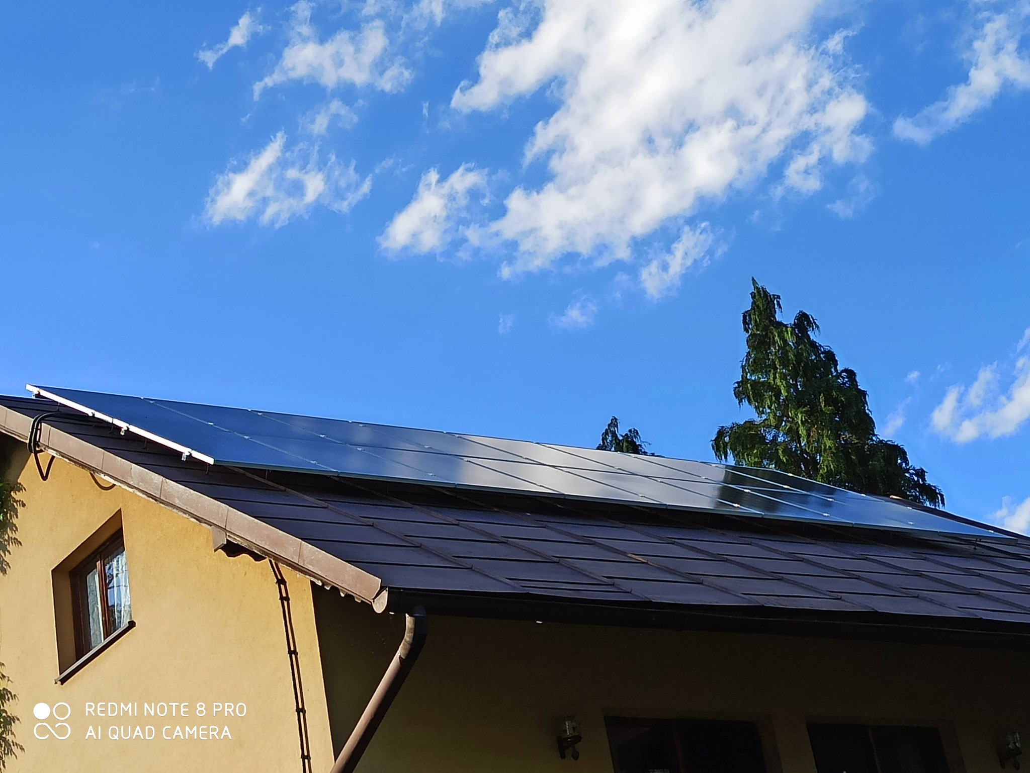 Panele słoneczne na brązowym dachu domu jednorodzinnego, błękitne niebo z chmurami w tle, widok z dołu.