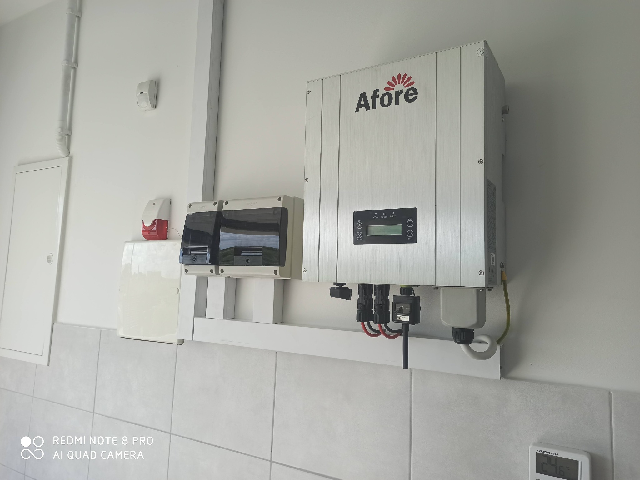 Szary falownik marki Afore zamontowany na białej ścianie z widocznymi przewodami i zabezpieczeniami, obok skrzynka rozdzielcza i czujnik alarmowy.