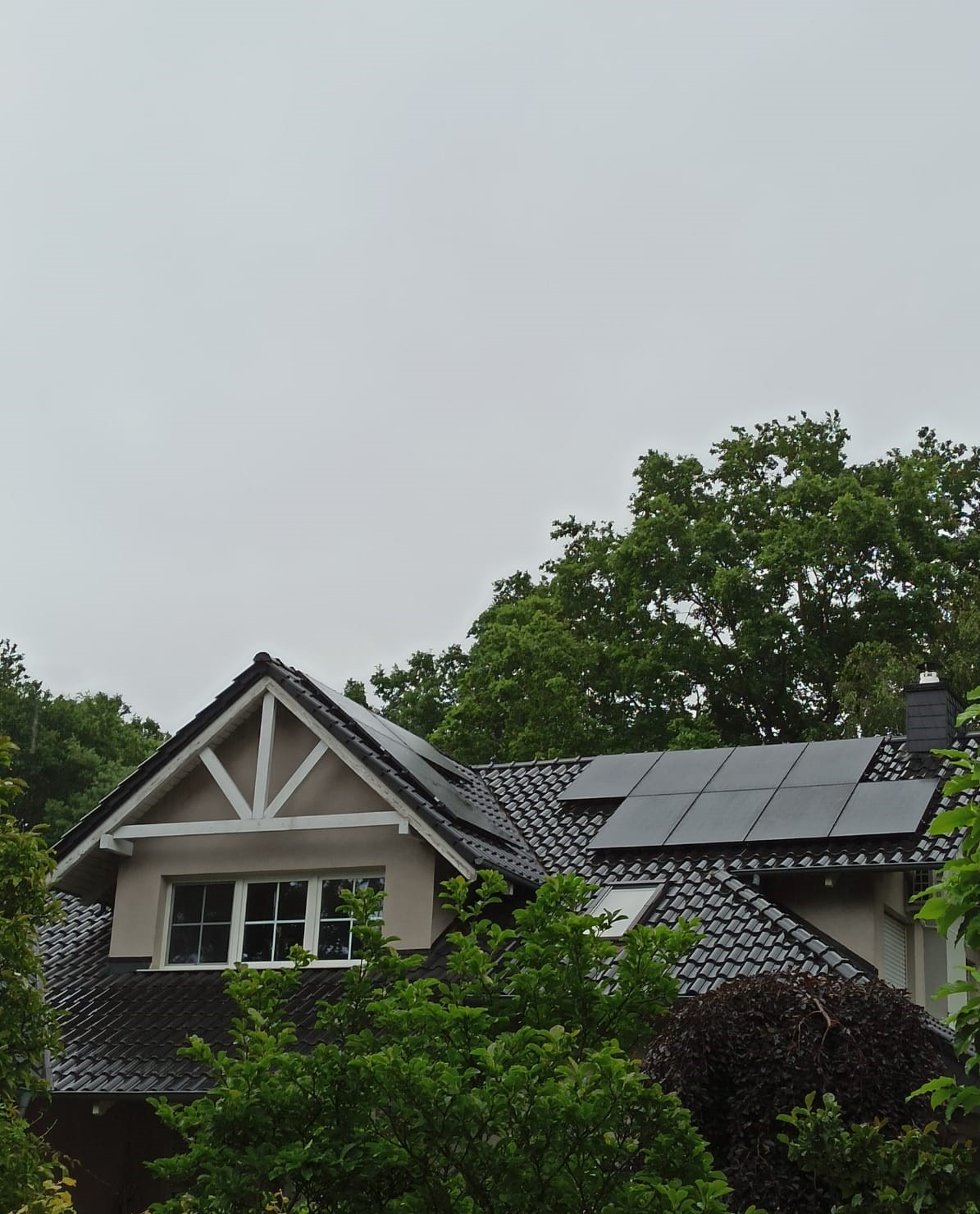 Częściowy widok domu z ciemną dachówką i panelami słonecznymi na dachu, widzianego zza drzew, pochmurne niebo.